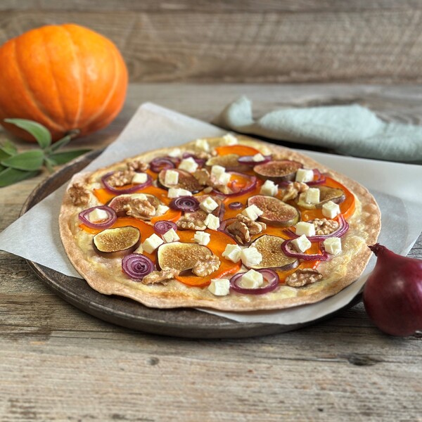 Flammkuchen mit Kürbisscheiben, roten Zwiebelringen, Feigen und Feta auf Backpapier auf einem Holzbrett angerichtet, im Hintergrund ein Kürbis, Salbei und ein graues Tuch