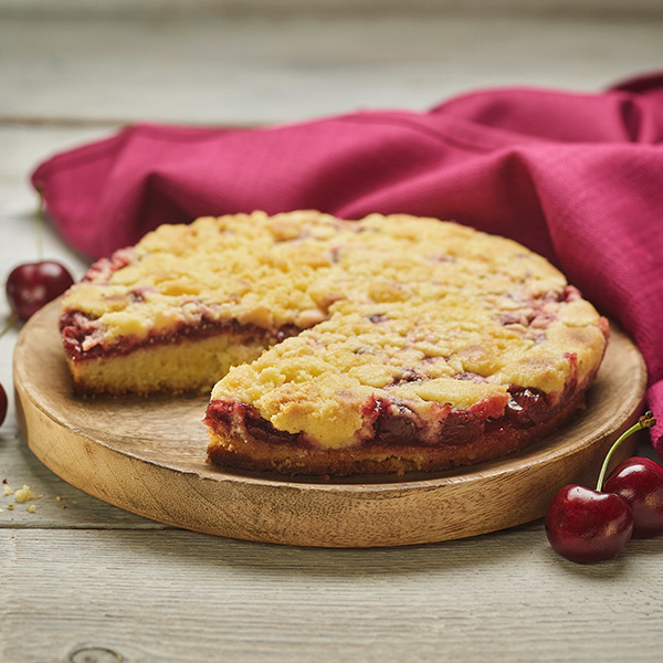Ein runder Kirsch-Streuselkuchen mit einem Stück, das herausgeschnitten wurde, steht auf einem Holzbrett, umgeben von frischen Kirschen und einem dunkelroten Tuch im Hintergrund.