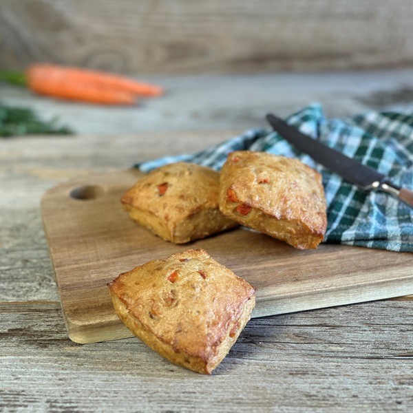 Drei Karottenbrötchen auf einem Holzbrett mit einem Messer und einem karierten Tuch im Hintergrund sowie zwei Karotten unscharf im Hintergrund